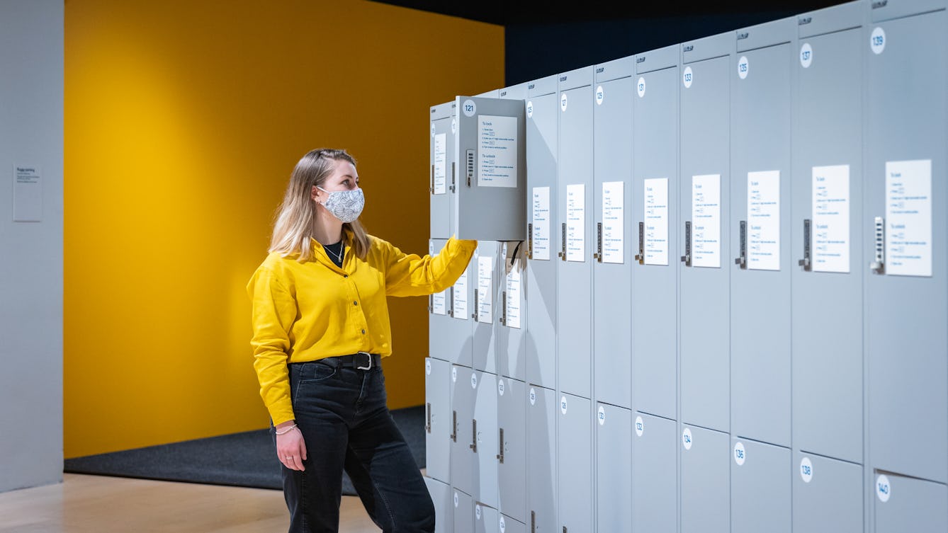 Photograph of a young woman wearing a face covering, a yellow shirt and black trousers, standing in front of a row of grey lockers.