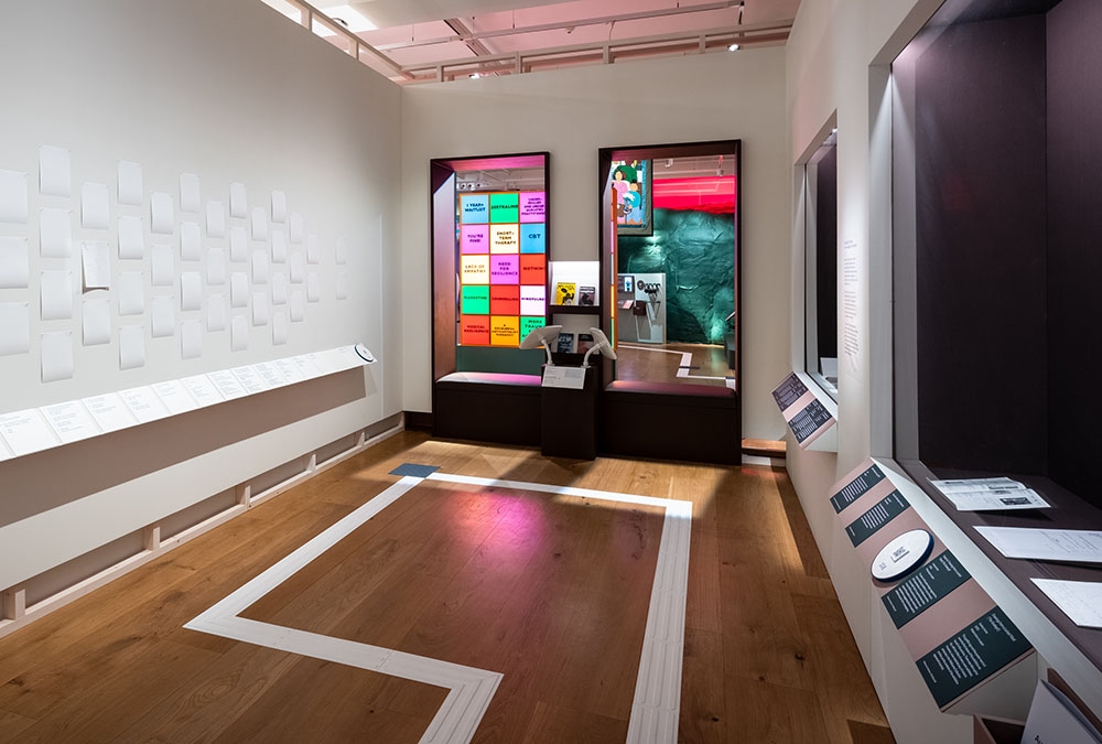 A gallery space in the exhibition called 'Audrey Amiss: the surviving exhibitions' showing items in a glass fronted display on the right, and small white cards attached ot the wall on the left. The back wall looks through to the accompanying exhibition, 'Rudy Loewe'.