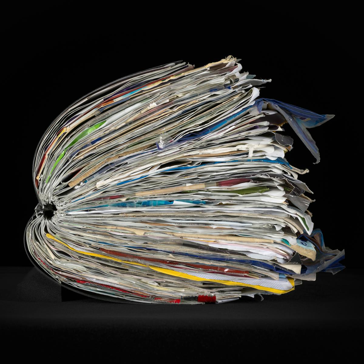 Side view of a large ring-bound scrapbook on a black background.  The scrapbook is bulging with ephemera pasted within it.  