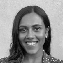 Black and white photograph of the head and shoulders of a young South Asian woman with long black hair. She is smiling and looking directly at the camera.