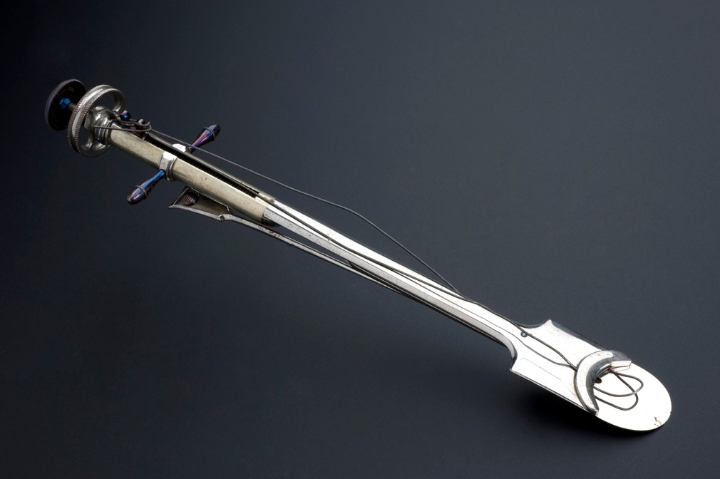 Colour photograph of a surgical implement. It has a silver-coloured device with a long rounded handle and a rounded-shovel-shaped end. There is a cable attached to a "u" shaped device on this end part and running up to a plunger on the handle at the other end.