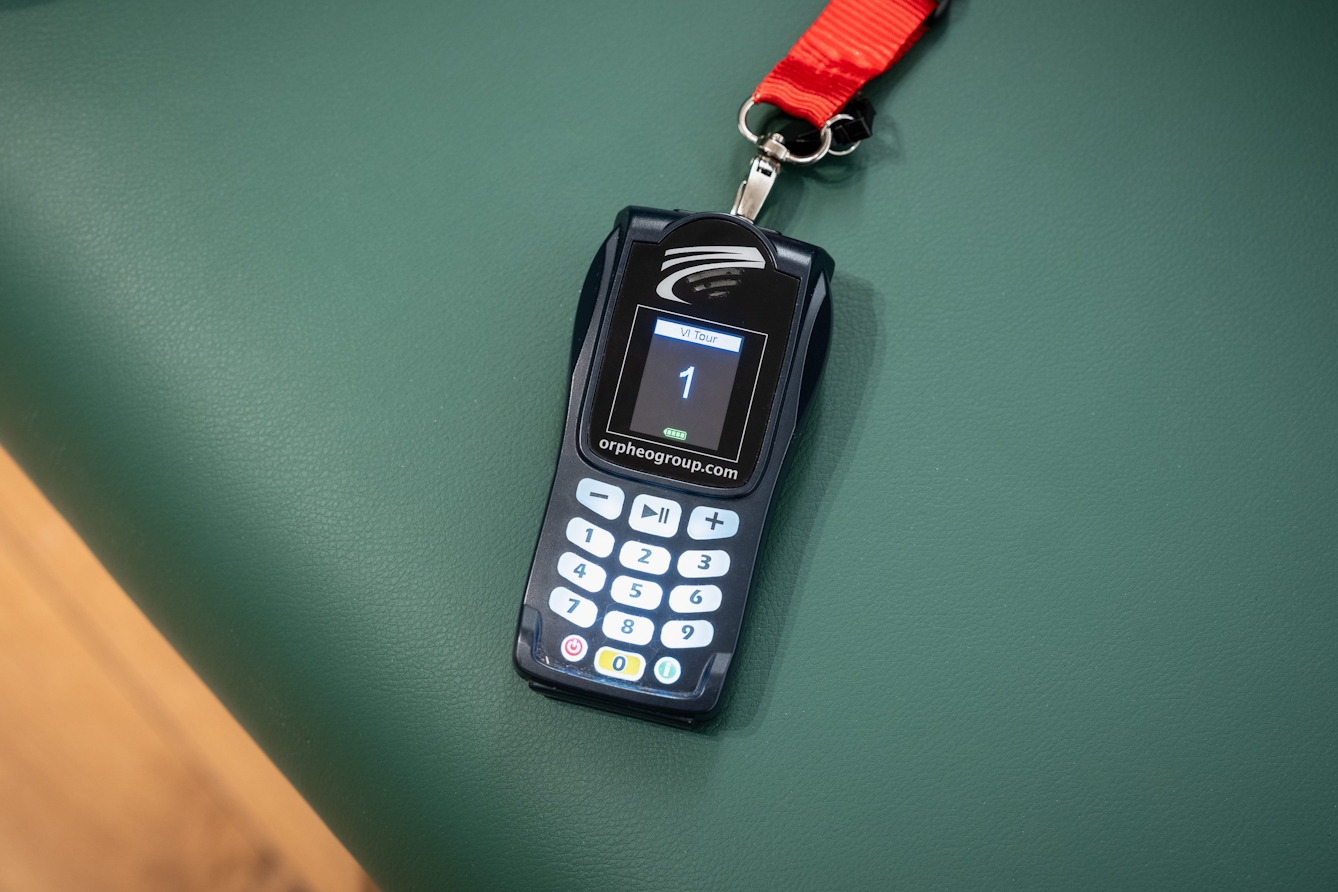Handheld audio guide device with a keypad and screen, resting on a green bench.
