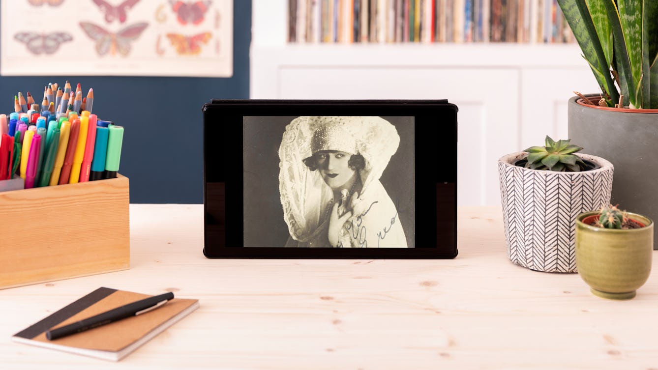A tablet standing on a desk. A black and white photograph on the screen shows Bert Errol, in character, wearing an elaborate 1920s white dress