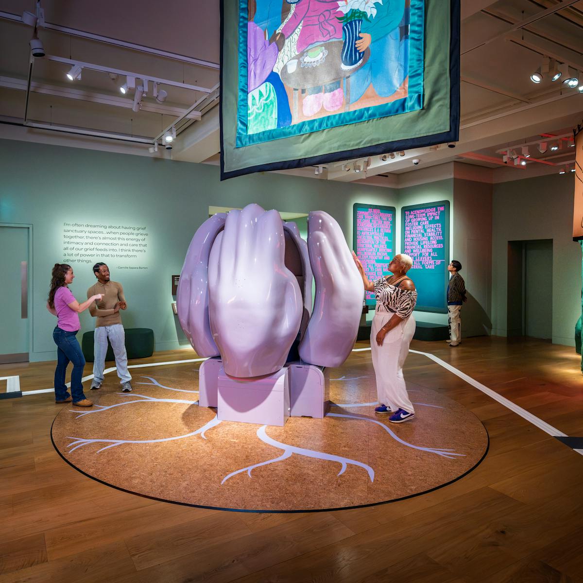 Photograph of four gallery visitors exploring a colourful exhibition space filled with artworks which center around a large sculpture of four vertical cupped hands. 