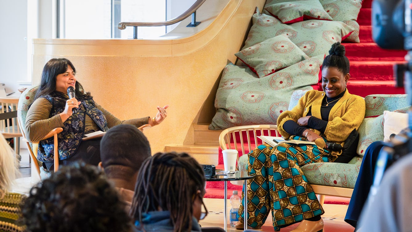 Two people sit together in a relaxed discussion space. The person on the left, holds a microphone and gestures while speaking.
