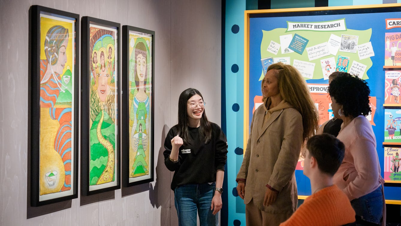 A Visitor Experience Facilitator standing in a brightly lit gallery giving a tour of the exhibition to visitors. There are large colourful artworks on the wall.