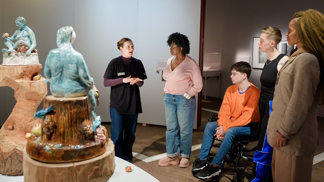 A Visitor Experience Facilitator using hand gestures while giving a tour of the exhibition. The group are looking at large ceramic sculptures on wooden plinths.