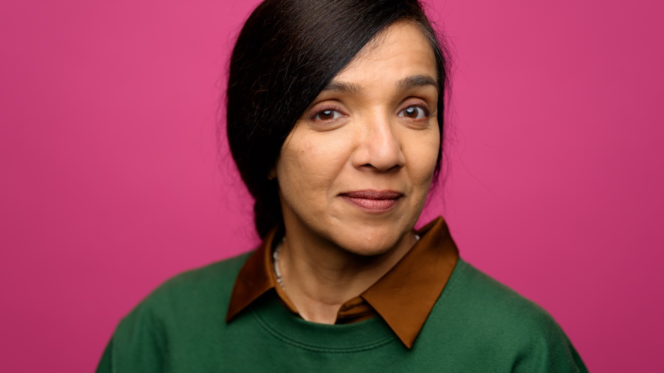 A close-up portrait of Rubbena Aurangzeb-Tariq looking straight to camera with a warm open gaze. The green of her jumper compliments the rose coloured background.