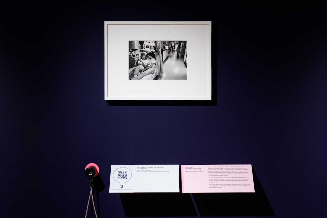 Photograph of a dark blue exhibition gallery wall showing a framed black and white photograph in a white frame. The photograph shows a hospital ward, with two people in casual clothes lying together on a narrow hospital bed surrounded by medical equipment, drinks and personal items, including photos and cards on the wall. Below the print is a caption panel and an audio guide ear cup.