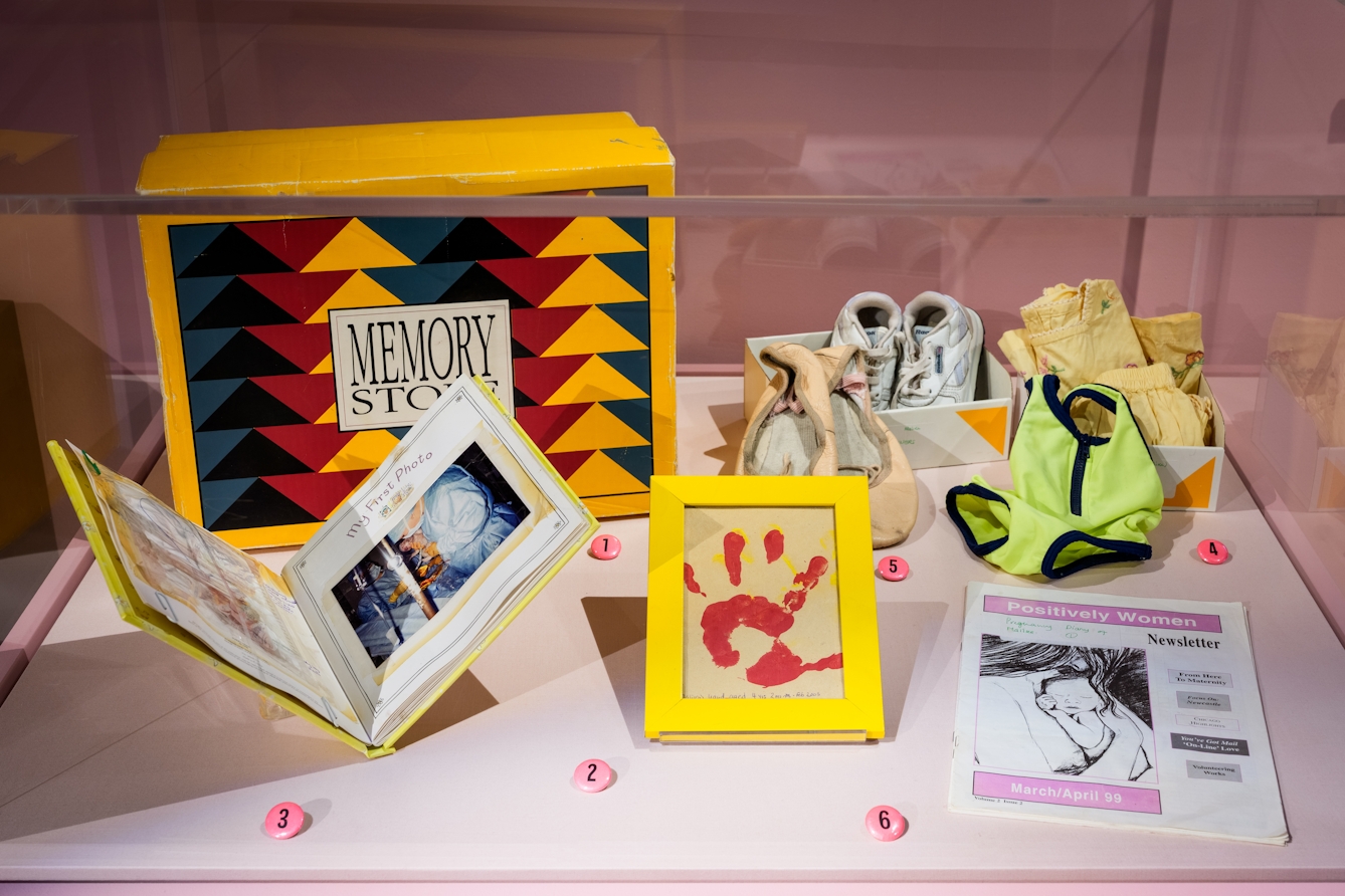 Photograph of an exhibition display case containing multiple items, including an open book, a framed hand print, a newsletter and ballet shoes.