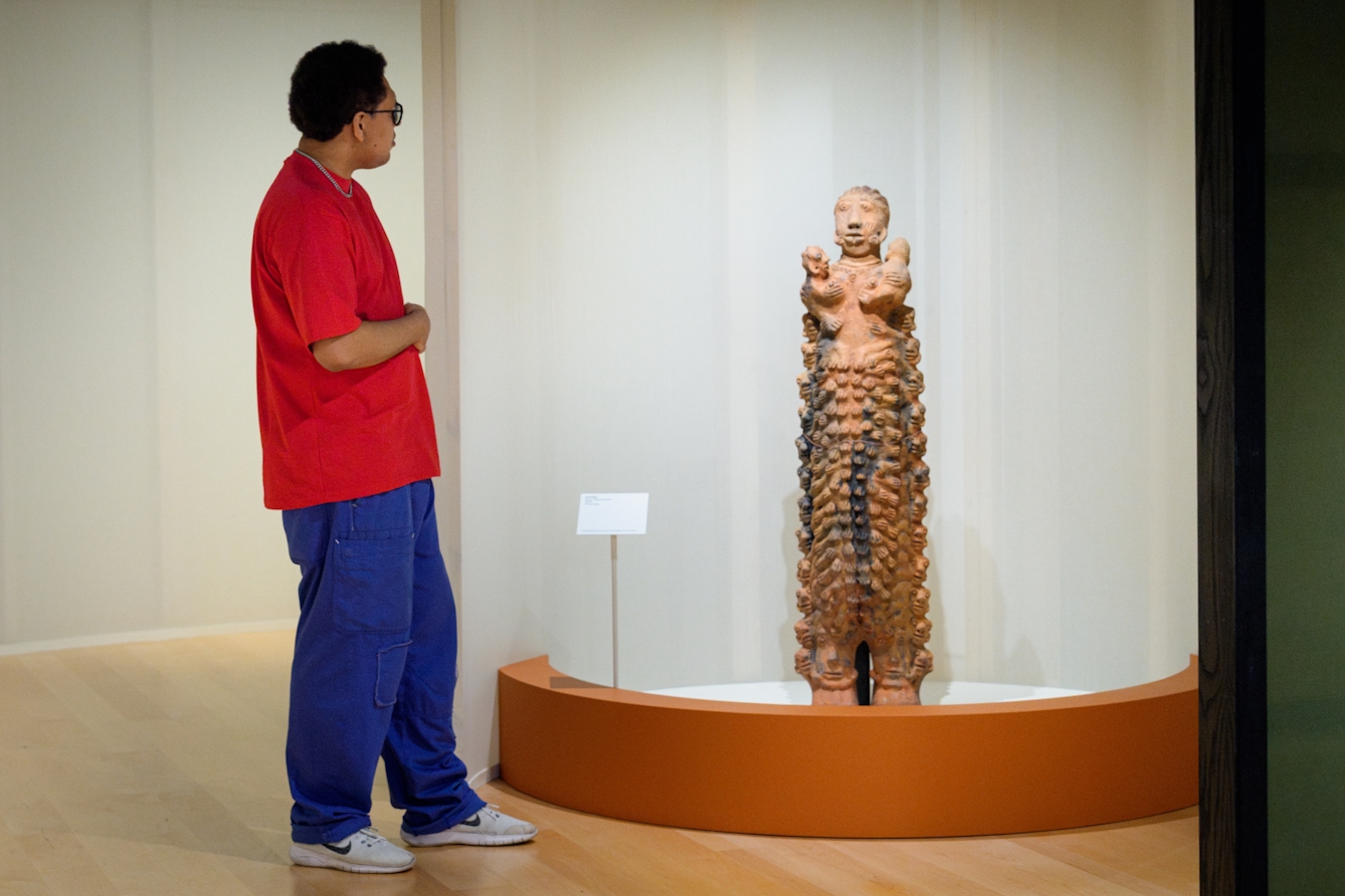 A gallery visitor looking at a tall clay scuplture of a woman, decorated with depictions of hands. 