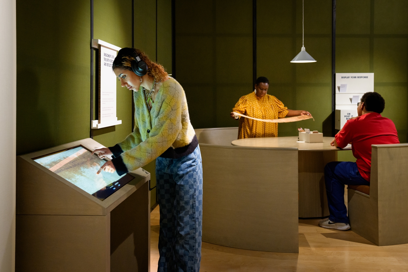 A gallery visitor using and interactive screen, in the background another visitor is holding a facsimile of a birthing scroll. 