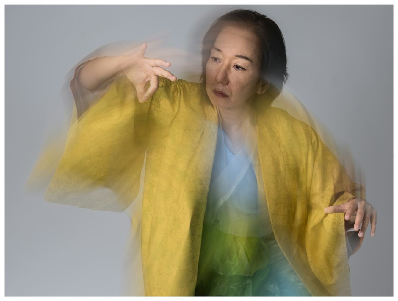 A portrait of Chisato Minamimura wearing a flowing yellow robe gesturing gracefully with blurred motion, set against a neutral background. The image conveys elegance and fluidity as she intensely studies her movement.