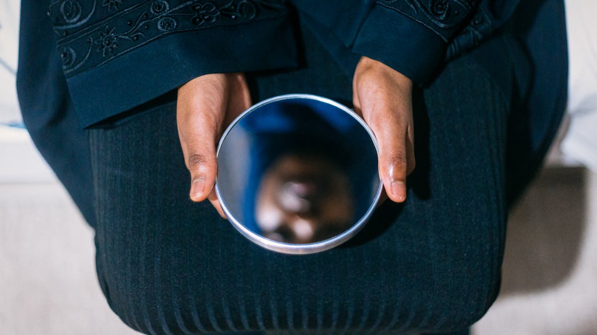 Photograph of a woman holding a circular mirror on her lap with both hands. In the reflection you can see her face, out of focus.