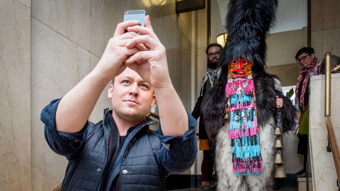 Photograph at a late night museum event. A man holds up a mobile phone, taking a photo over his shoulder of the scene behind him. The scene is a person descending a staircase dressed in a fur outfit complete with mask and a fur covered headdress.