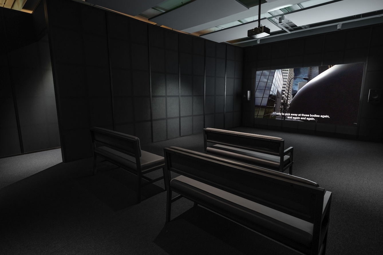 Photograph of a dark exhibition space featuring a projected video with subtitles on a large screen. 3 bench seats are arranged facing the screen. The walls of the room are criss-cross of black wooden battens on grey fabric. The video shows a computer generated film of a glass and steel building being engulfed by a large black expanding spherical mass.