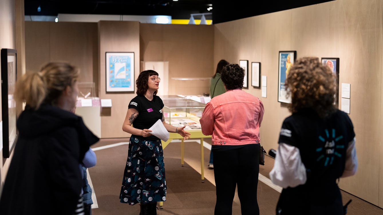 A Visitor Experience Assistant standing in a brightly lit gallery giving a tour of the exhibition to visitors.