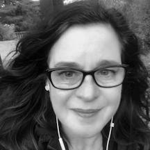 Black and white, head shot of a woman with long dark hair, wearing spectacles and trailing leads from earbuds, against the background of a park.