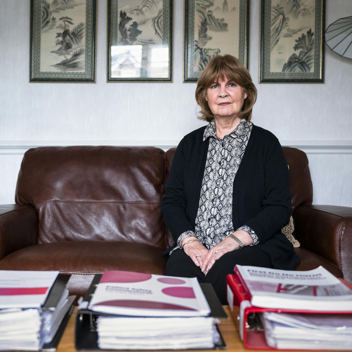 Photographic portrait of Marie Lyon sat on her brown leather sofa in her living room. She is looking straight to camera with a strong, serious expression. In front of her on the coffee table are 2 full lever arch files and printed reports.