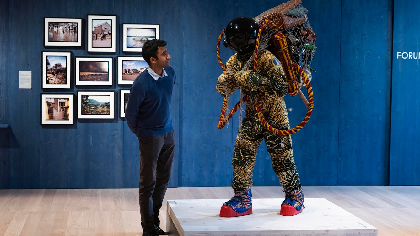 Photograph of an exhibition gallery space, with a blue stained wood wall in the background. In the foreground is a life-size artwork of a figure resembling an astronaut, carrying a large net containing assorted objects including a suitcase.