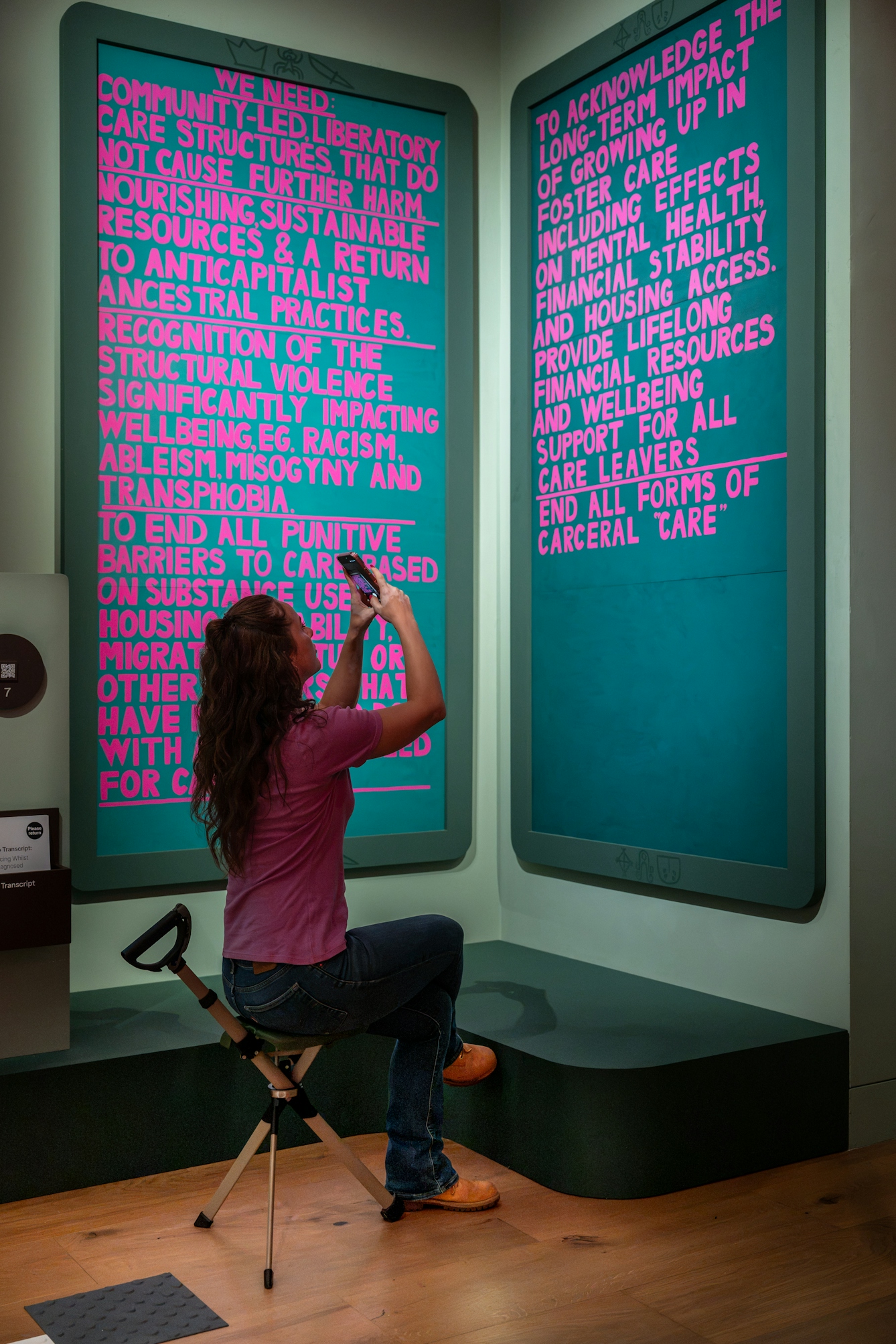 Photograph of a person sitting down and taking a photograph of two large text-based artworks with bright pink lettering on a dark background, displayed in a gallery corner.