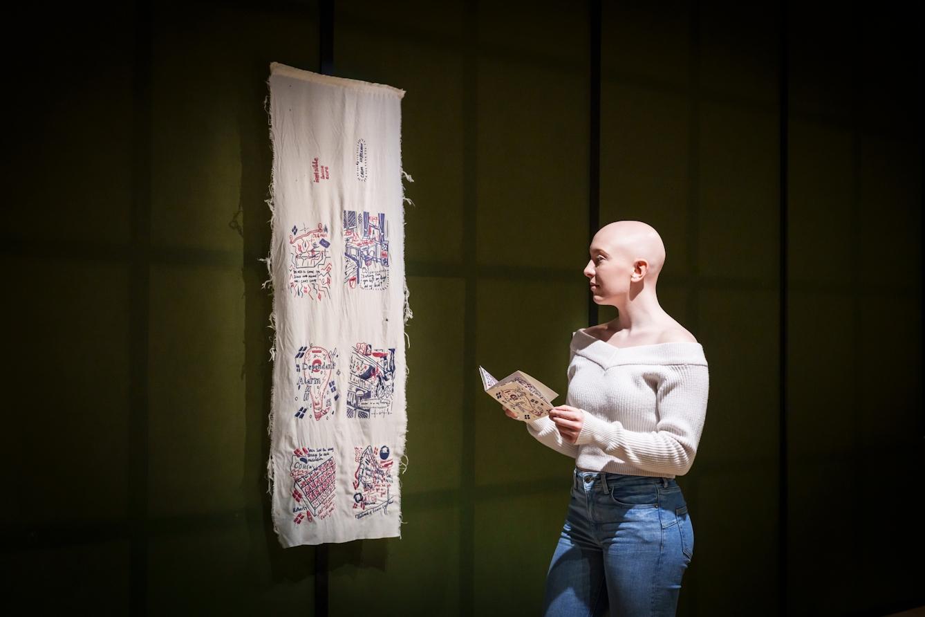 Photograph of a gallery visitor standing in front of a hanging fabric artwork made up of drawings and text stitched with thread. In their hands they are holding a corresponding paper zine of the artwork.