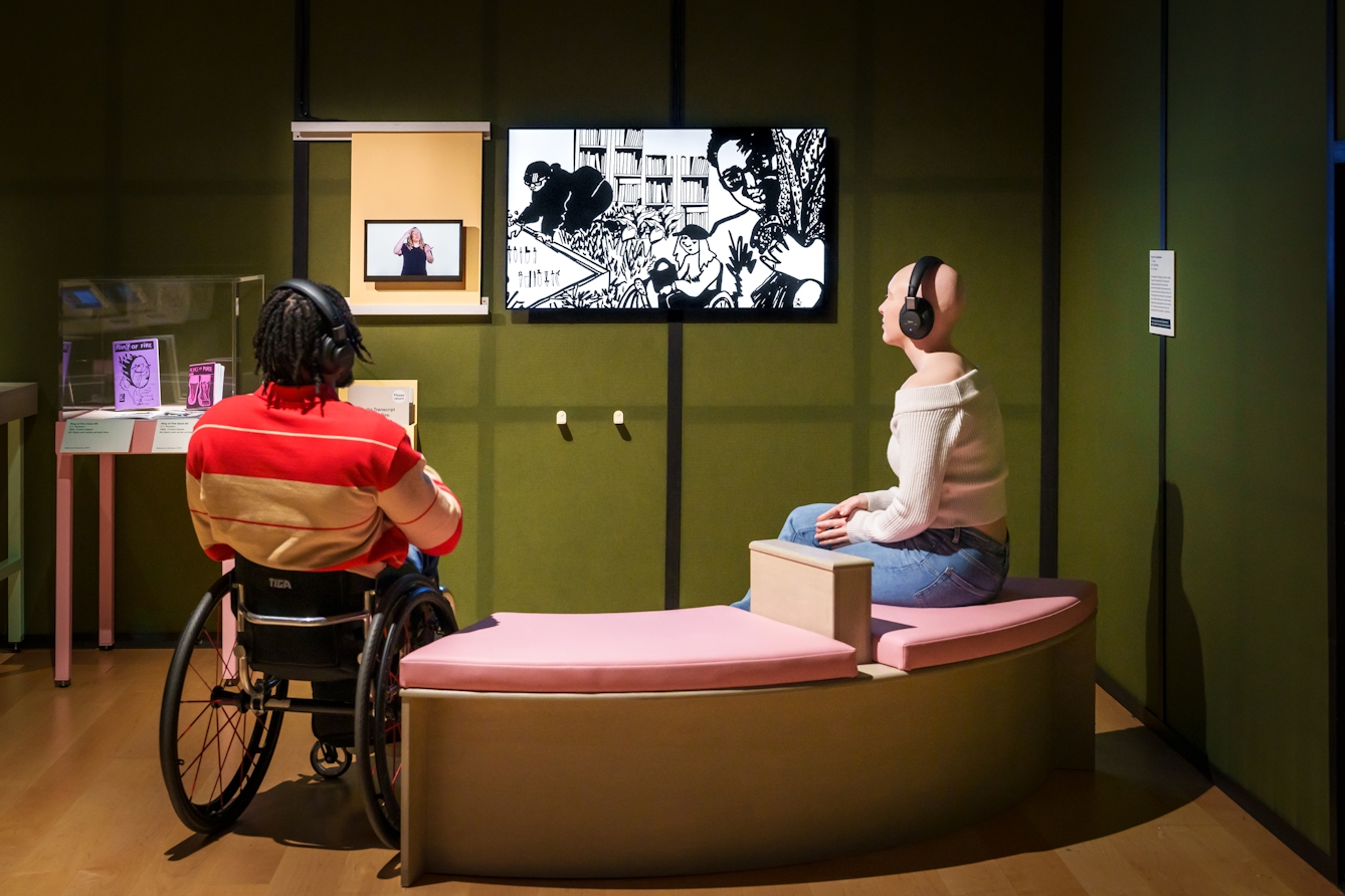 Photograph of two gallery visitors sat watching a film with headphones. Next to the film, a smaller screen shows a BSL translation.