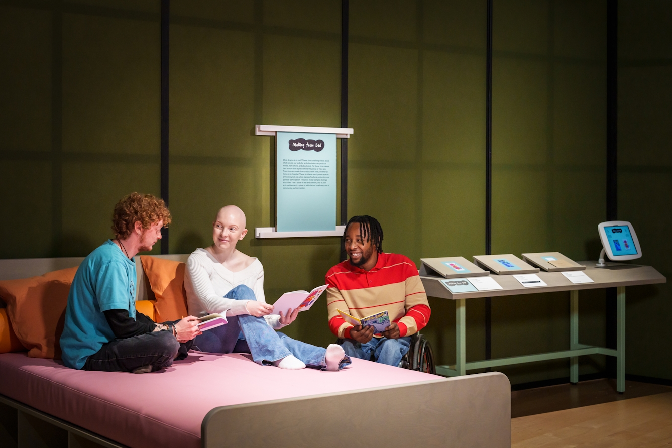 Photograph of three people chatting in an exhibition gallery. They each have a zine held open in their hands. Two of them are sat on a large double bed, made up of a pink mattress and orange pillows, the other sits beside them, next to the bed in their wheelchair. In the background is a display table with more zines and a tablet in a holder.