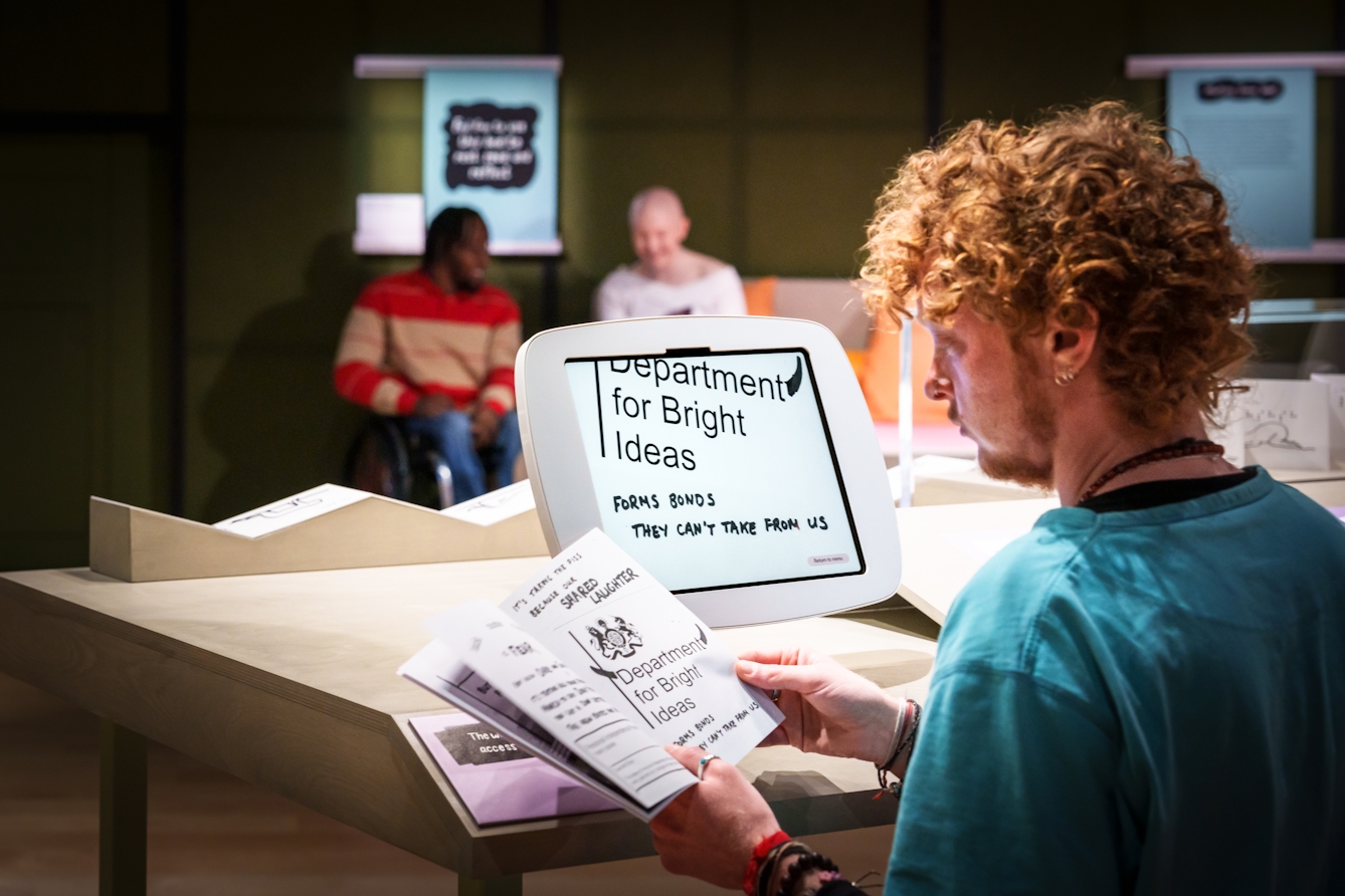 A young man sits at a table reading a booklet which he holds in his hands.  A monitor on the table in front of him shows an enlargement of what he sees in the booklet.