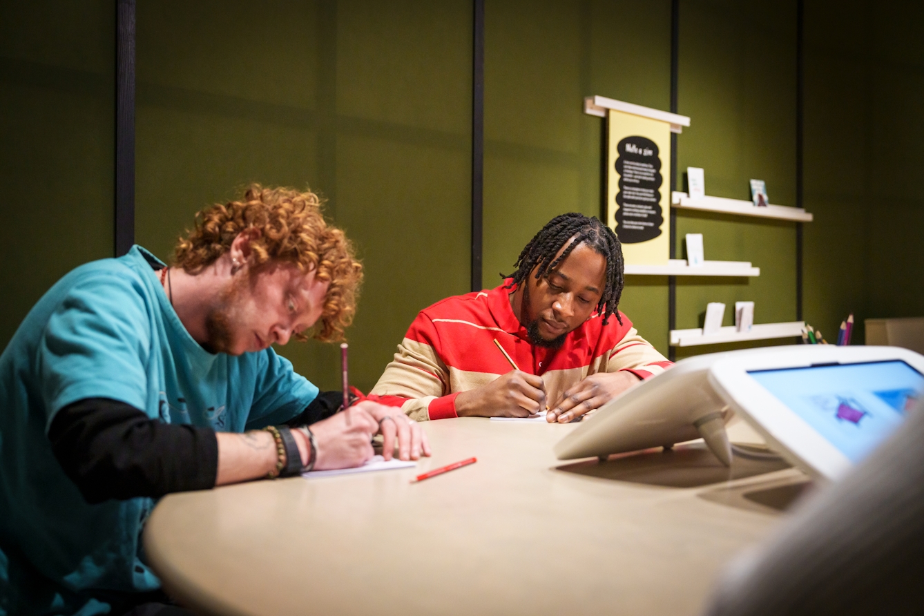 Photograph of two gallery visitors creating a personal zine, at a large round table. Each has a pencil in hand and is deep in concentration.