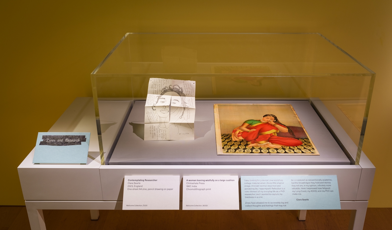 Photograph of a gallery display case containing two paper works. One is a handmade folded zine showing a hand drawn face and handwriting. The other is a colour drawing of a woman leaning wistfully on a large cushion. 