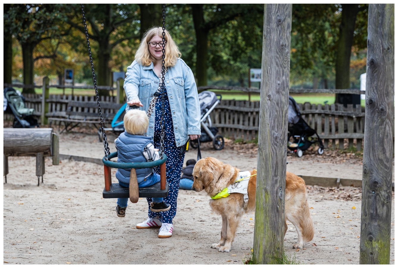Photograph of Amy in a kids playground pushing her small child on the swings. They are in mid conversation while her assistance dog looks on.