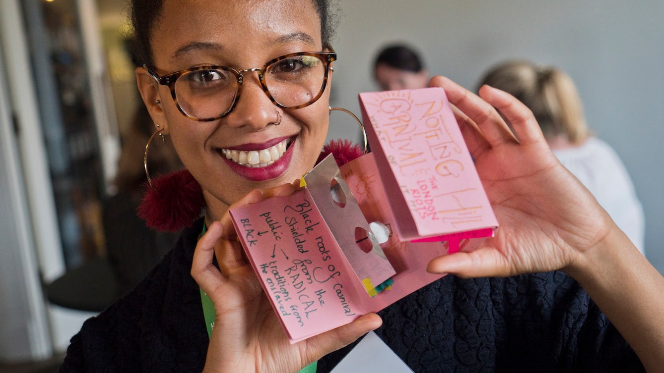 Photograph of a young woman with large round glasses and dark hair, tied back. She is smiling and looking straight at the camera while holding up a small folded paper document of a zine that she has made.