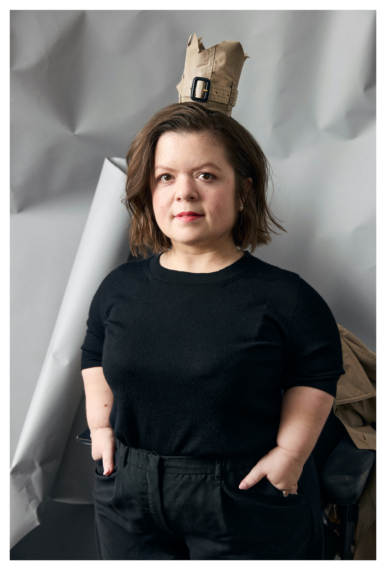 Photographic portrait of Sinéad, dressed in black, hands in pockets. She is looking straight to camera with a hint of a smile. Standing out against the grey crumples backdrop is the crown on her head made from the cuff of a Burberry trenchcoat.