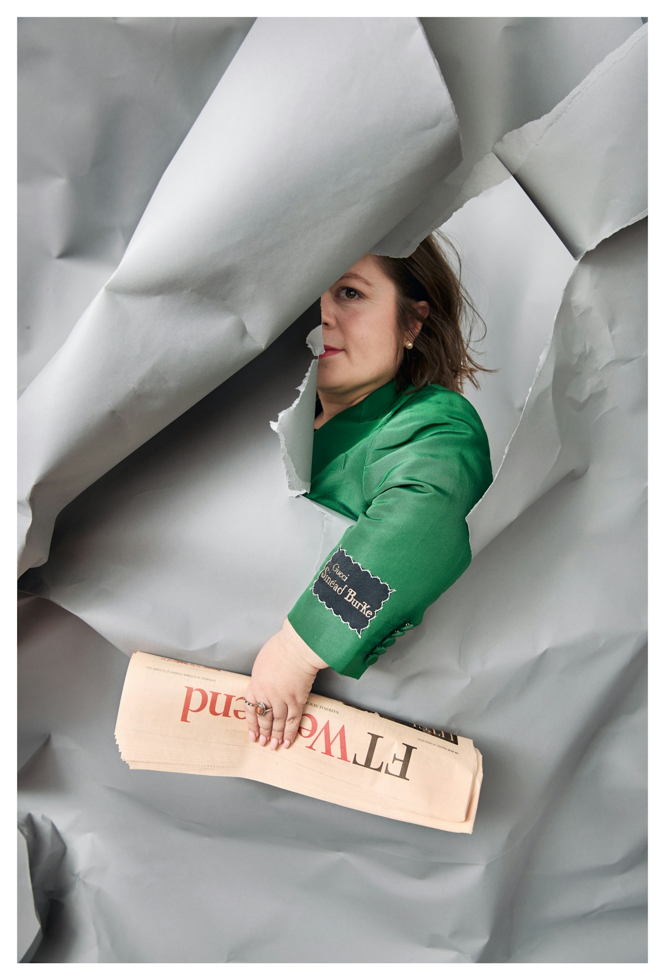 Photograph of Sinéad through a hole roughly torn in the grey paper background. Her arm pokes through the hole, clutching a folder FT Weekend newspaper. Half of her Face is just visible through the hole and on the cuff of the emerald green jacket she is wearing, is a label which reads, "Gucci. Sinéad Burke".